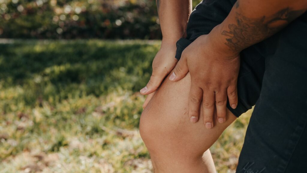 pexels-photo-7298631-7298631 A close-up image of a person holding their knee, indicating pain or injury, with greenery in the background.